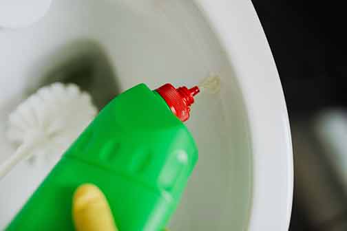 A person pouring liquid drain cleaner into a white toilet bowl while holding a brush.